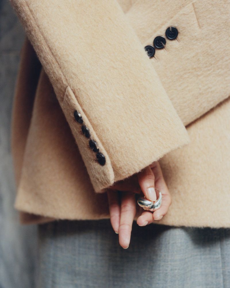 Close-up detail of a model wearing a textured beige wool blazer with buttoned cuffs and holding a sculptural silver ring.
