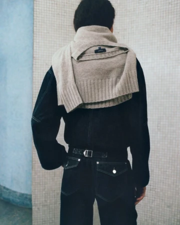 Model seen from behind wearing dark denim with contrast stitching and a beige ribbed knit sweater draped over the shoulders.