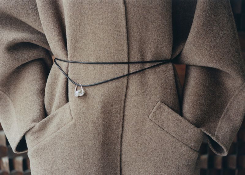 Woman's back in warm taupe wool coat tied with black cord and delicate silver pendant against abstract brown background. 