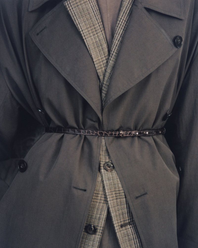 Close-up of a belted charcoal grey trench coat layered over a brown plaid blazer with dark buttons.