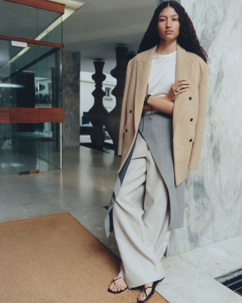 A stylish biracial woman in a beige coat and wide-leg trousers poses indoors against a marble wall, showcasing a blend of fashion and architecture.