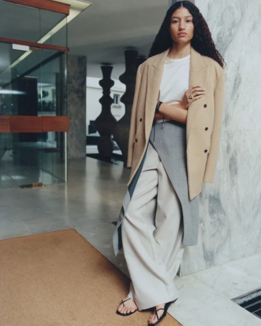 A stylish biracial woman in a beige coat and wide-leg trousers poses indoors against a marble wall, showcasing a blend of fashion and architecture.