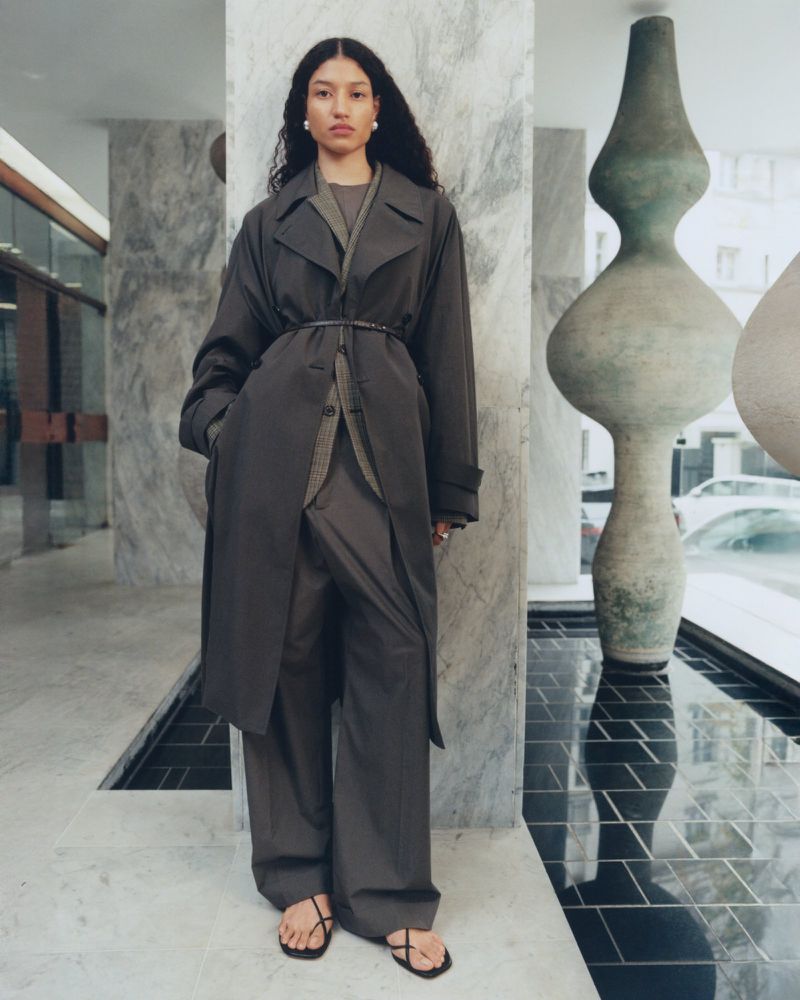 An adult female fashion model poses indoors wearing a dark trench coat, wide-leg pants, and strappy sandals in a fashion editorial shot.