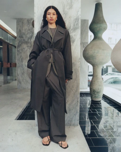 An adult female fashion model poses indoors wearing a dark trench coat, wide-leg pants, and strappy sandals in a fashion editorial shot.