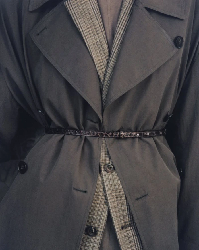 Close-up of a belted charcoal grey trench coat layered over a brown plaid blazer with dark buttons.