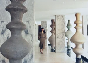 Model Keny Salgado wearing a long taupe belted coat walking past large ceramic sculptures and marble columns.