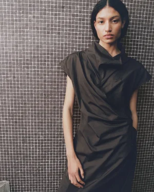 Model Keny Salgado wears a dark draped sleeveless dress while posing in front of a dark mosaic tiled wall.