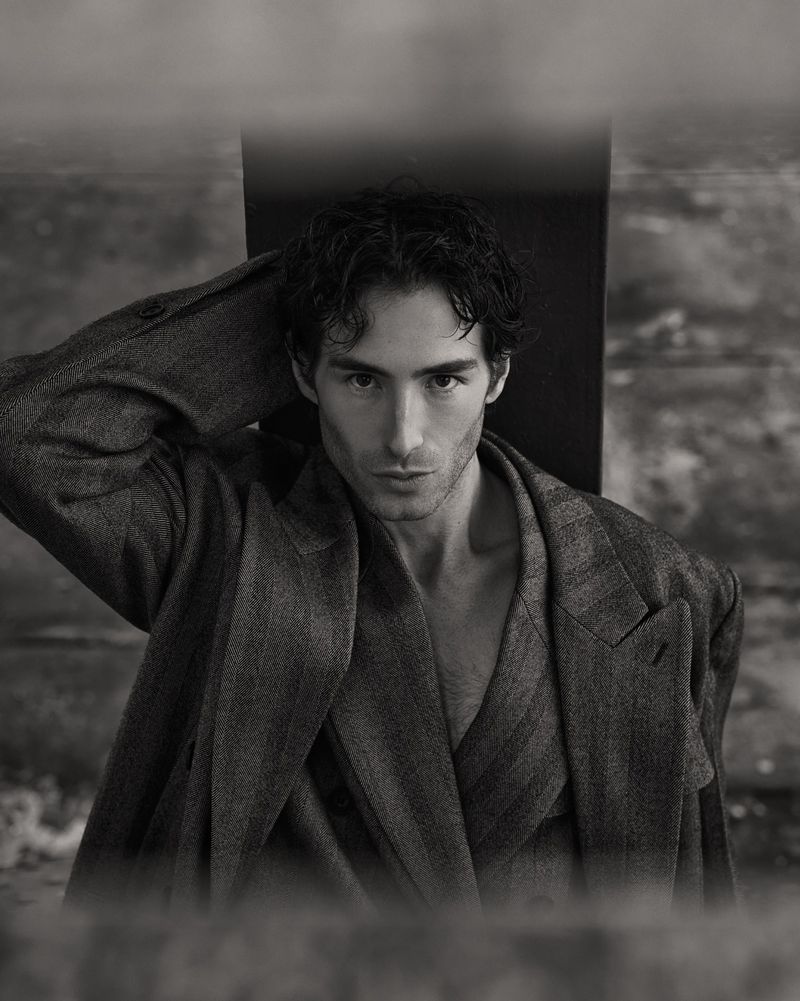 Monochrome portrait of a handsome adult man with dark hair wearing an open suit jacket, gazing directly at the viewer indoors.