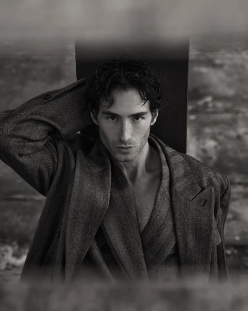 Monochrome portrait of a handsome adult man with dark hair wearing an open suit jacket, gazing directly at the viewer indoors.