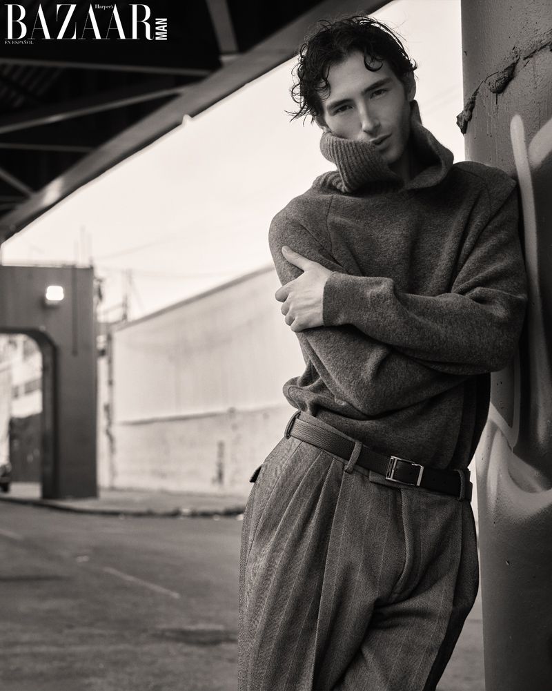 Monochrome editorial portrait of a man in a high-neck knit sweater and pleated trousers leaning against a post on an urban street.