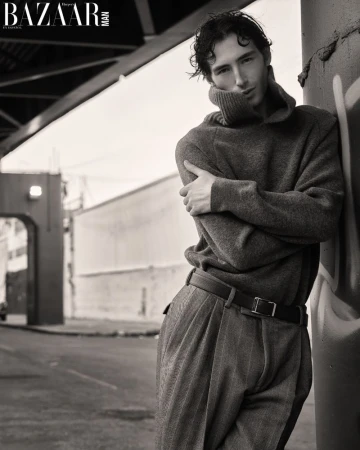 Monochrome editorial portrait of a man in a high-neck knit sweater and pleated trousers leaning against a post on an urban street.