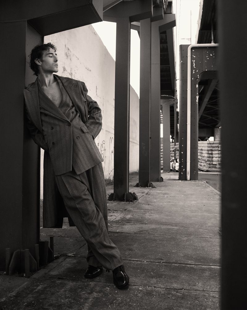 A man in a suit leans against a column outdoors. The image is a black and white portrait of a suited man posing in an urban area.
