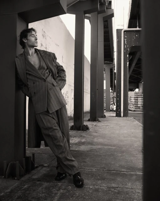 A man in a suit leans against a column outdoors. The image is a black and white portrait of a suited man posing in an urban area.