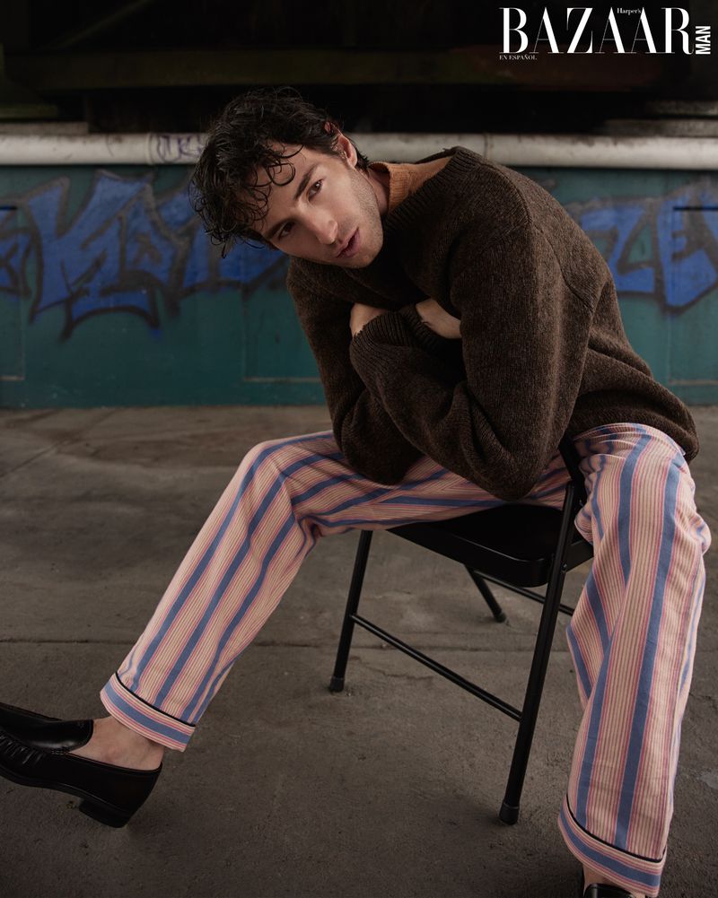Man in brown sweater and striped pants poses seated against a graffiti wall in a fashion shot for Bazaar Man magazine.