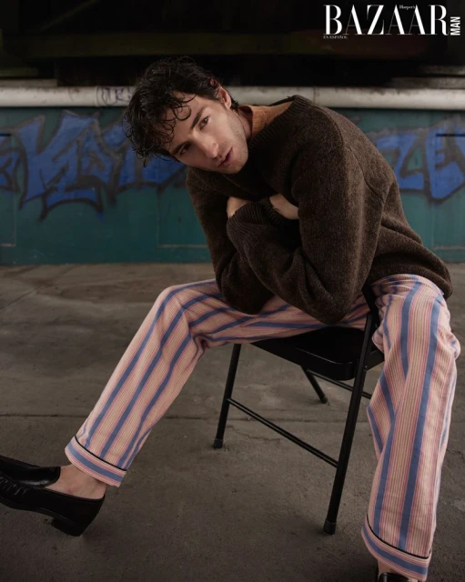 Man in brown sweater and striped pants poses seated against a graffiti wall in a fashion shot for Bazaar Man magazine.