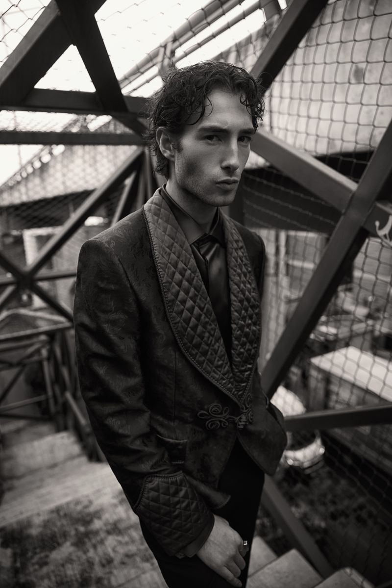 Elegant man poses on stairs in a tailored blazer and dress pants, with a neutral facial expression, captured in a black-and-white photograph.