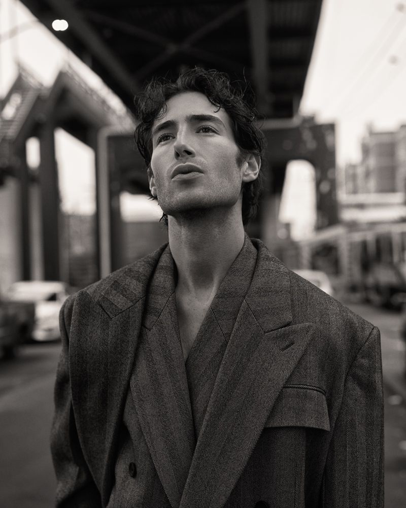 Monochrome portrait of a man wearing a suit jacket, looking upward with buildings and a bridge in the blurred background.