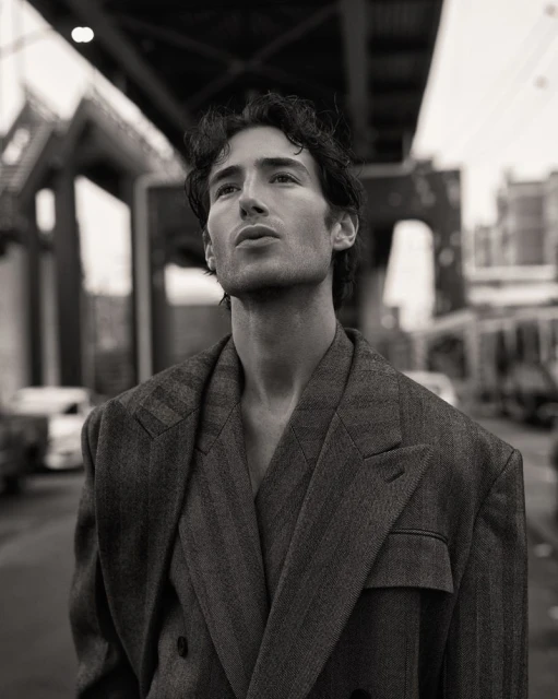 Monochrome portrait of a man wearing a suit jacket, looking upward with buildings and a bridge in the blurred background.