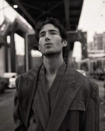 Monochrome portrait of a man wearing a suit jacket, looking upward with buildings and a bridge in the blurred background.