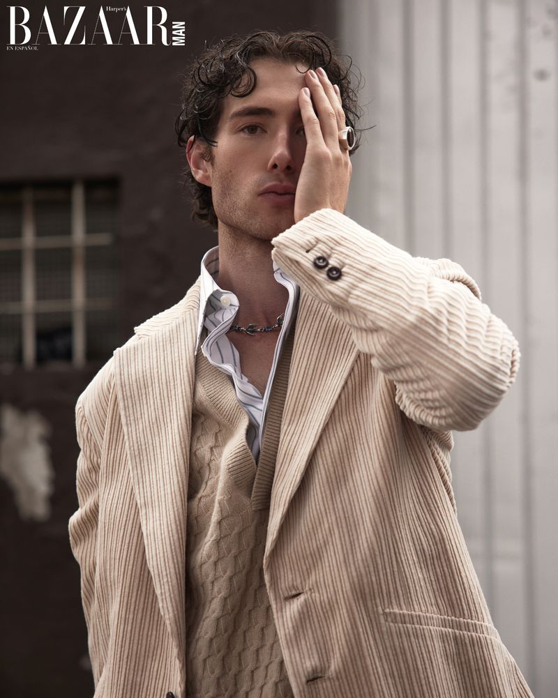 Man with curly hair wearing a beige corduroy jacket and cable knit sweater holding his hand over his face