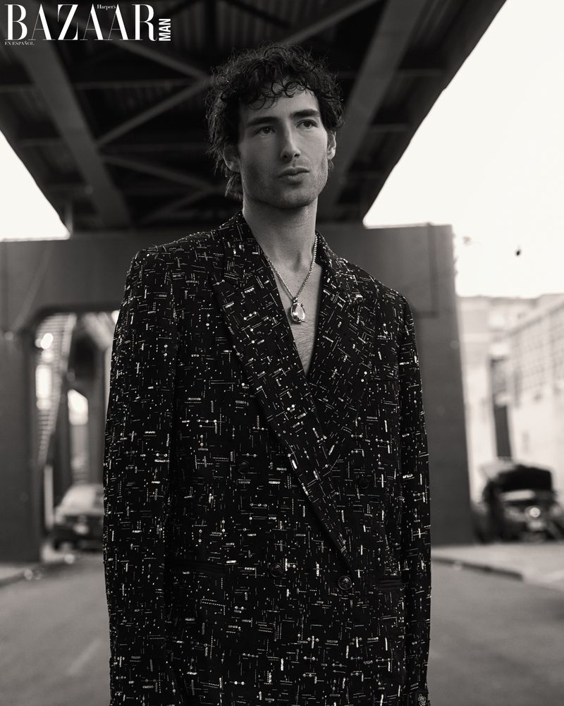 Monochrome fashion portrait of a man in a patterned blazer wearing a pendant necklace beneath an overpass bridge.