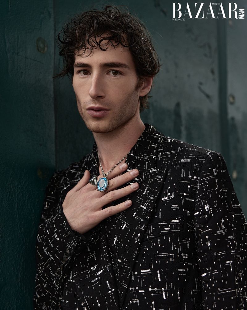 Portrait of a dark-haired man in a patterned blazer with a large blue ring and necklace, posing indoors with a serious expression.