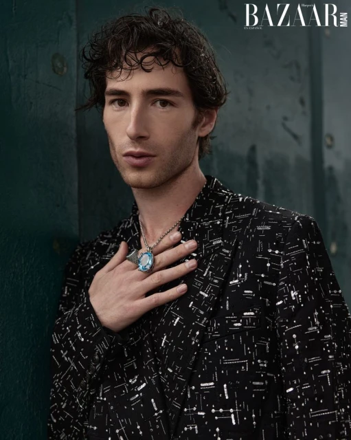 Portrait of a dark-haired man in a patterned blazer with a large blue ring and necklace, posing indoors with a serious expression.