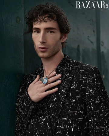 Portrait of a dark-haired man in a patterned blazer with a large blue ring and necklace, posing indoors with a serious expression.