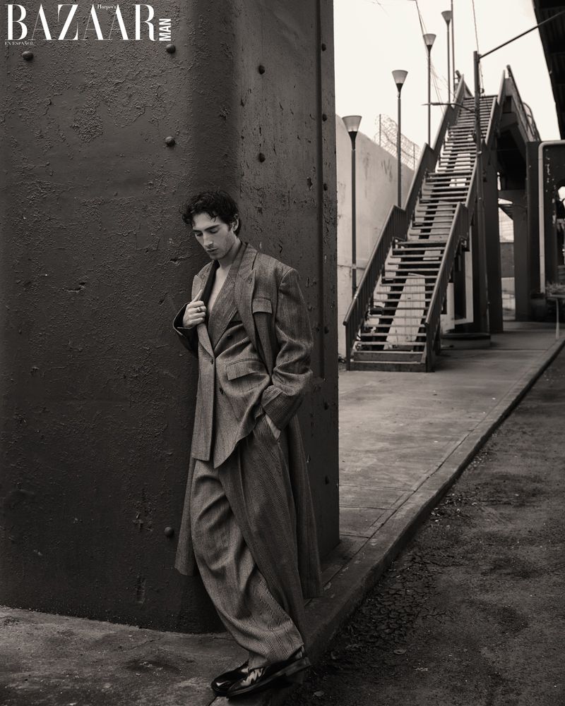 Man in tailored suit posing against a steel column, with staircase and urban elements visible. Fashion editorial shot in monochrome.