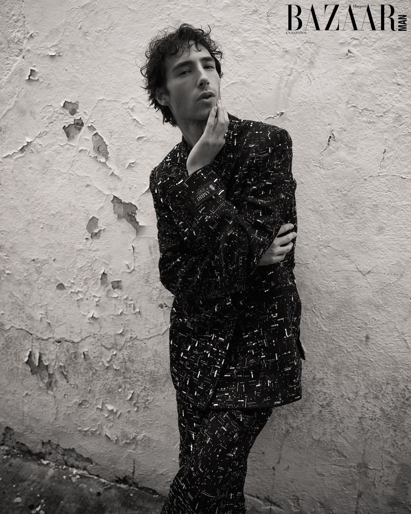 Monochrome portrait of a young adult man in patterned suit leaning against peeling wall for Bazaar Man magazine cover.