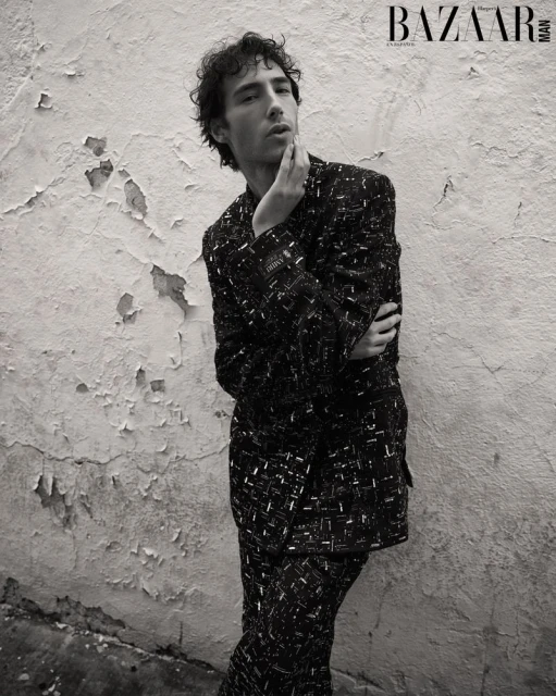 Monochrome portrait of a young adult man in patterned suit leaning against peeling wall for Bazaar Man magazine cover.