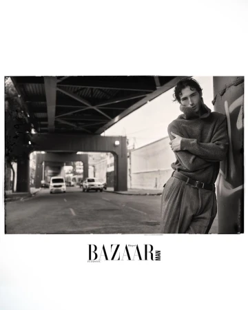 Black and white fashion shot of a male model leaning against a wall under an overpass wearing a sweater and pants, photographed at eye-level.