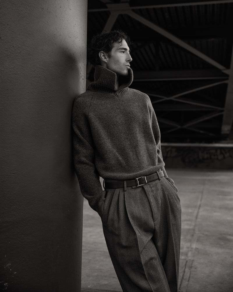Monochrome portrait of a young adult male in a wool sweater and pleated pants, leaning against a column with a somber expression.