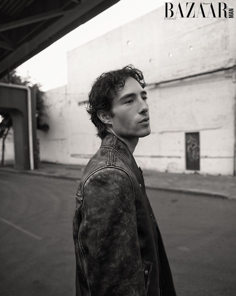 Monochrome portrait of a man with dark hair wearing a textured leather jacket, set against an urban background with an underpass.