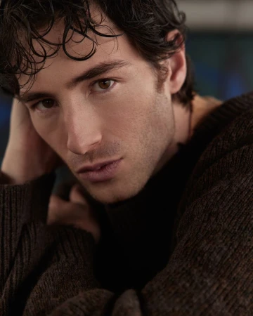 Portrait of a young man with dark hair and brown eyes, wearing a brown sweater, posing indoors with a blurred background.