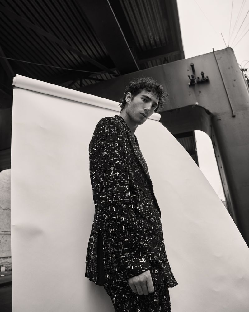 Black-and-white fashion shot: a young adult male poses, wearing a patterned blazer and pants, against a white backdrop with industrial features.