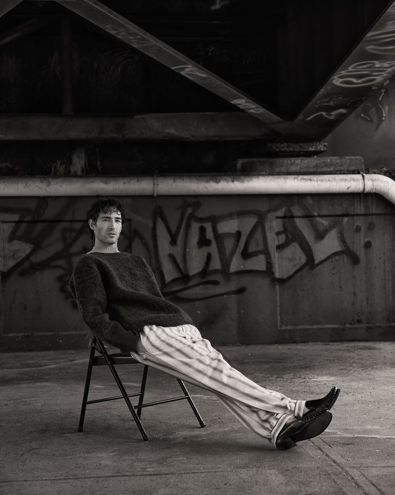 Black and white portrait of a young adult male in a sweater and striped pants, sitting on a folding chair beneath a bridge with graffiti.