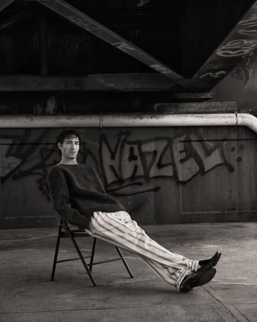 Black and white portrait of a young adult male in a sweater and striped pants, sitting on a folding chair beneath a bridge with graffiti.