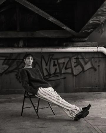 Black and white portrait of a young adult male in a sweater and striped pants, sitting on a folding chair beneath a bridge with graffiti.