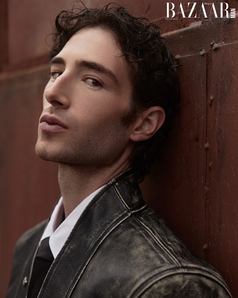Close-up portrait of a handsome adult male with dark wavy hair wearing a jacket, a white shirt, and a dark tie leaning against a wall.