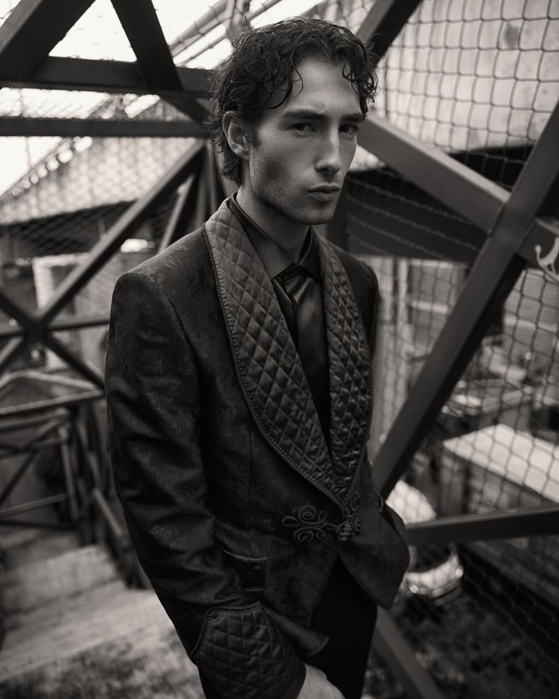 Monochrome portrait of a male model wearing a quilted velvet suit, posing in front of a geometric metal structure, neutral expression.