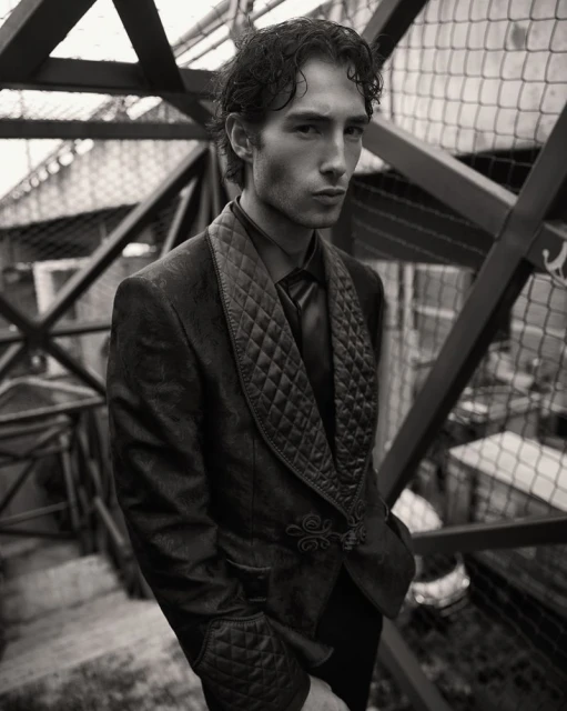 Monochrome portrait of a male model wearing a quilted velvet suit, posing in front of a geometric metal structure, neutral expression.
