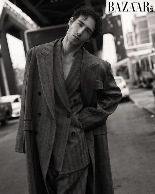 Monochrome portrait of a slender man in a striped suit and overcoat stands on a city street next to parked cars and an elevated train.