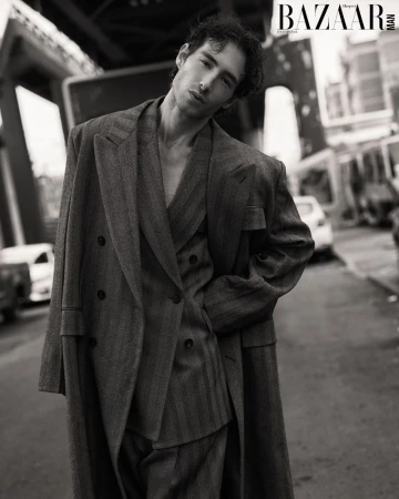 Monochrome portrait of a slender man in a striped suit and overcoat stands on a city street next to parked cars and an elevated train.