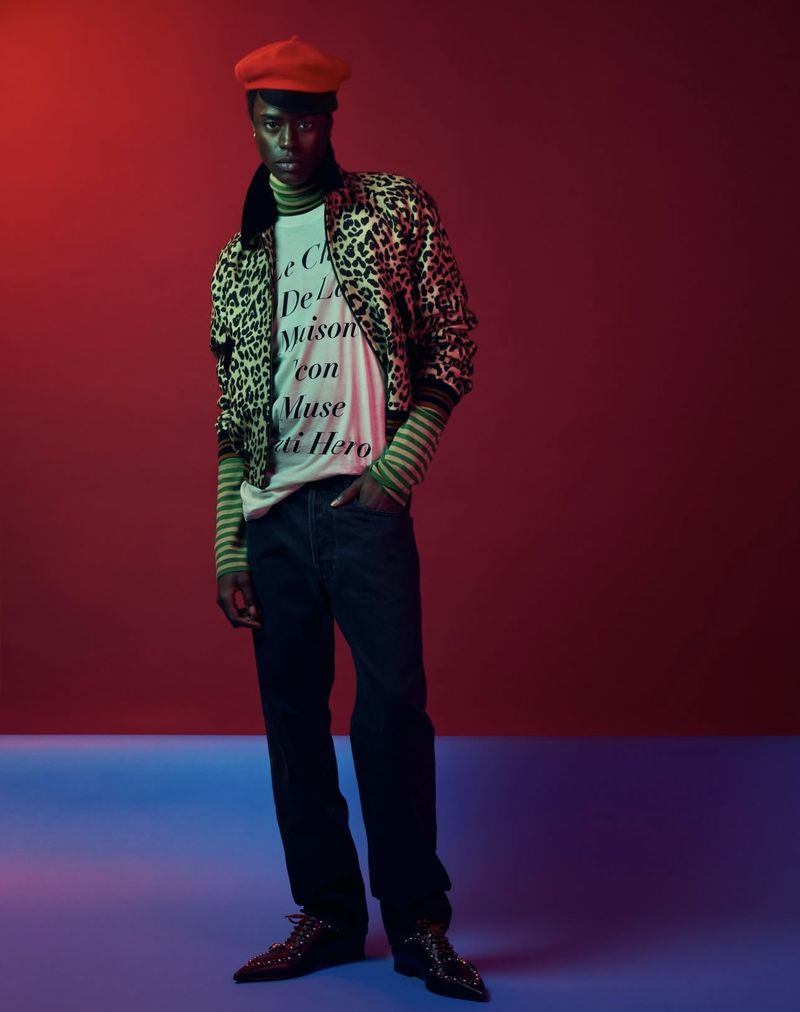 Fashion portrait of a young adult Black male wearing a patterned jacket, tee shirt, jeans, beret, and pointed shoes against a red background.