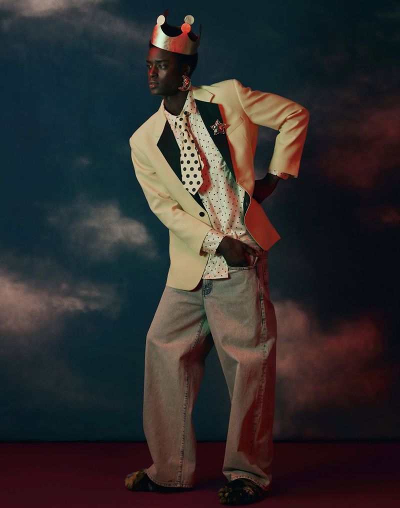 Full-body studio shot of a Black male model in a cream suit jacket, polka dot shirt, baggy jeans, and a crown, posing in a colorful studio.
