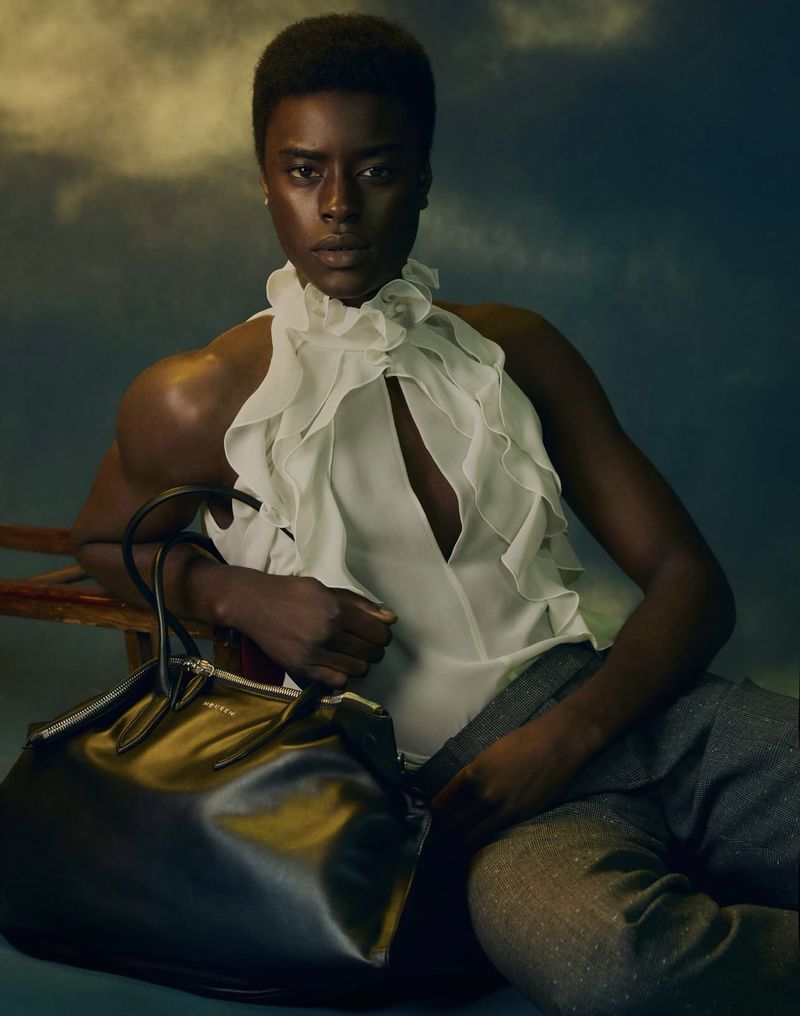 Elegant African-American woman in white ruffled blouse seated with a black leather handbag, studio portrait, fashion concept.