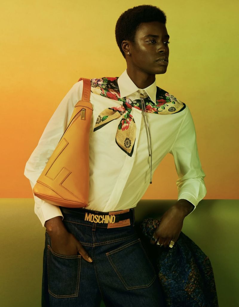 Fashion portrait of a black male model in a white shirt, Moschino belt, jeans, and shoulder bag against a yellow and green background.