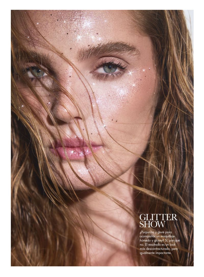 Close-up beauty shot of a woman with glitter makeup and brown hair, wearing a glossy lip, set against a blurred, neutral background.