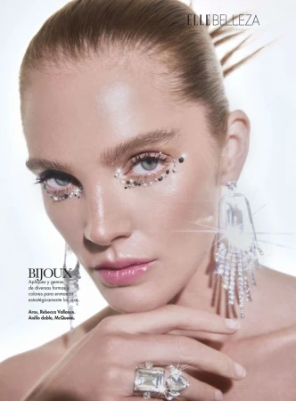 Close-up of a fair-skinned woman with glitter around her eyes, wearing statement earrings and ring, posing in a beauty context.